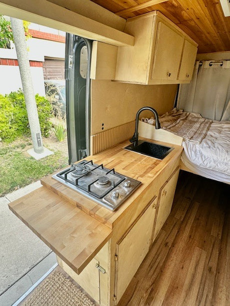 Compact RV rental kitchen & living space. Features a wood countertop with sink, 2-burner stove, pull-out table, & cozy bed area.