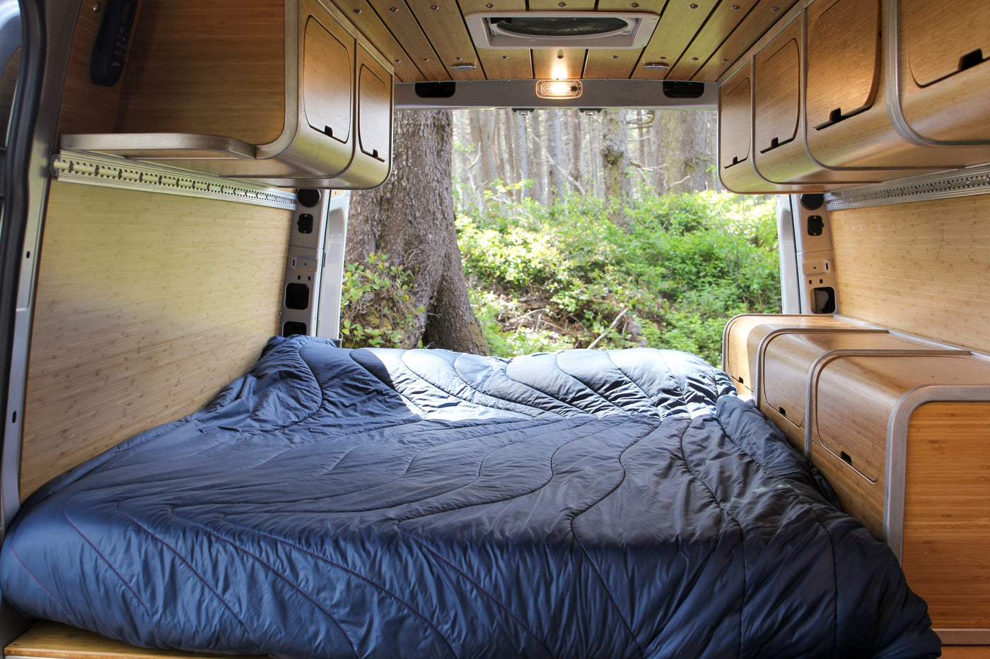 This RV rental offers a cozy, well-appointed interior with a large bed, bamboo accents, and a picturesque forest backdrop. — photo 7