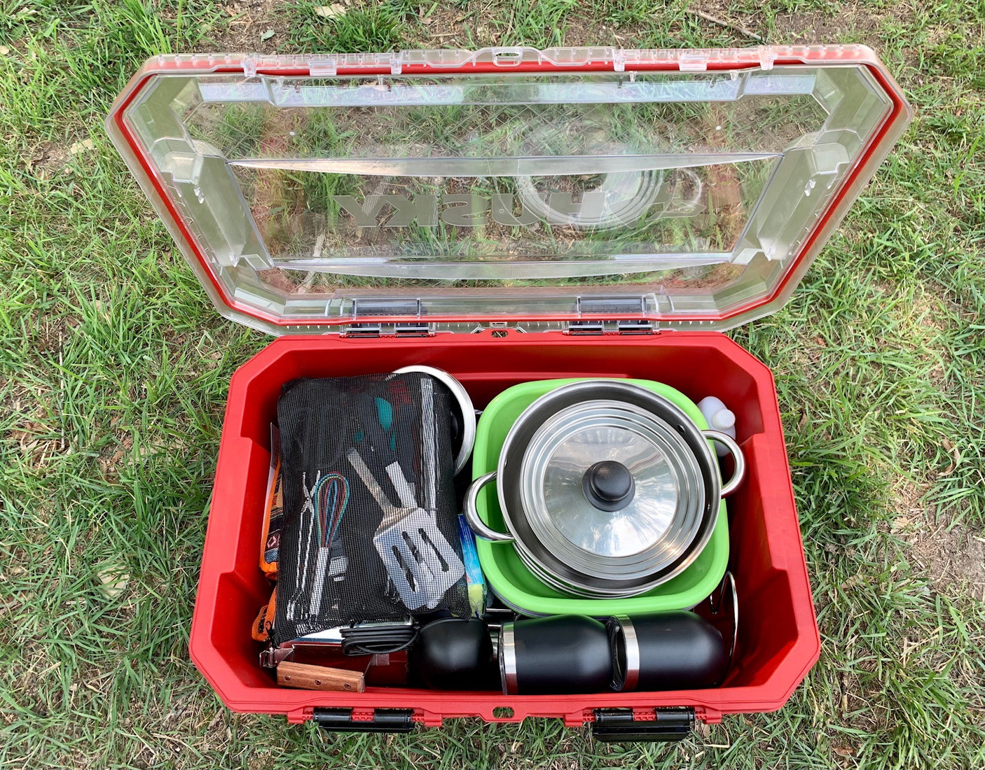 A red outdoor case open on grass, revealing neatly packed camping cooking gear: pots, utensils, and mugs. Ideal for your RV rental adventure!