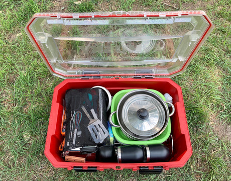 A red outdoor case open on grass, revealing neatly packed camping cooking gear: pots, utensils, and mugs. Ideal for your RV rental adventure!