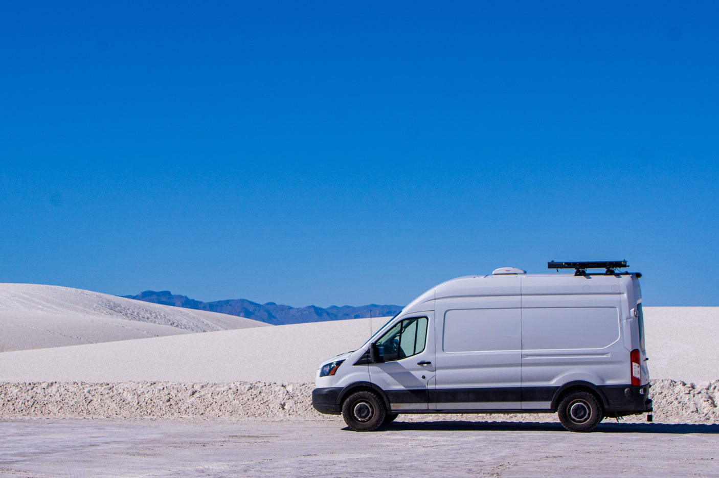 White RV rental van parked in a vast white sand dune desert under a clear blue sky, with distant mountains.