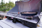 A Coleman Triton camp stove sizzles with food in a pan on a table, ideal for outdoor cooking on an RV rental trip.