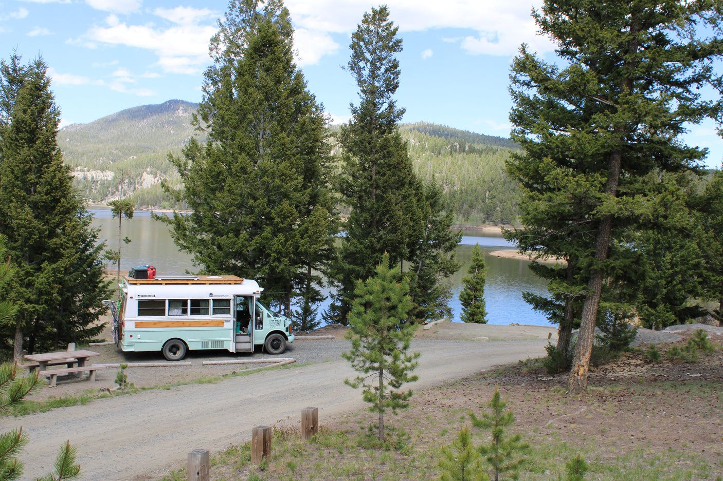Skoolie RV rental parked by a serene lake with tall pine trees and mountains in the background.