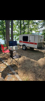 This lakeside RV rental features a white teardrop camper, camping chairs, and an outdoor TV set up on gravel. Enjoy glamping!