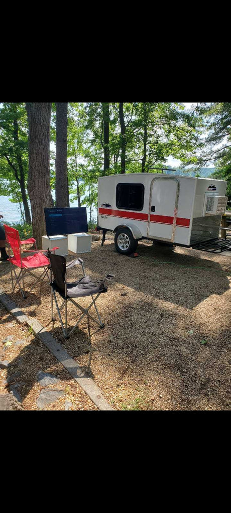 This lakeside RV rental features a white teardrop camper, camping chairs, and an outdoor TV set up on gravel. Enjoy glamping!