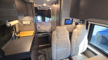 Modern rv rental interior with dark wood and light seats, featuring a kitchenette, rear bed, and control panel.