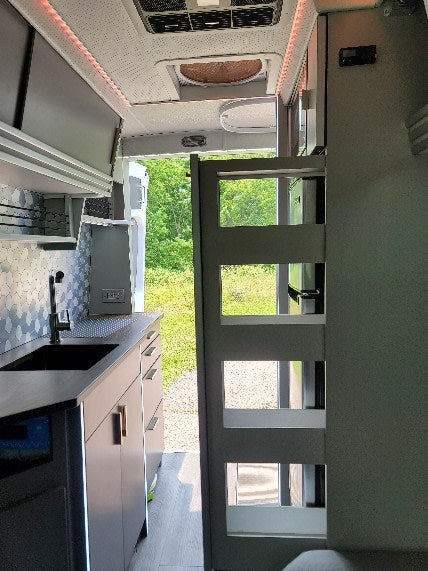 Inside a modern **rv rental**: compact kitchen with sink, gray cabinets, overhead storage, and open rear doors revealing a green outdoor view.