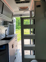 Inside a modern **rv rental**: compact kitchen with sink, gray cabinets, overhead storage, and open rear doors revealing a green outdoor view.