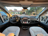 Inside an RV rental: a wide-angle view of the front cabin, showing the dashboard, steering wheel, and light-colored seats.