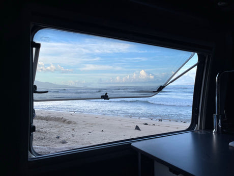 A view from inside an RV, through its open window, showing a serene sandy beach and ocean waves under a cloudy sky. Ideal for an RV rental getaway.