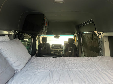 Inside an RV rental, a comfy bed fills the foreground, leading to the front cabin with seats and overhead storage.