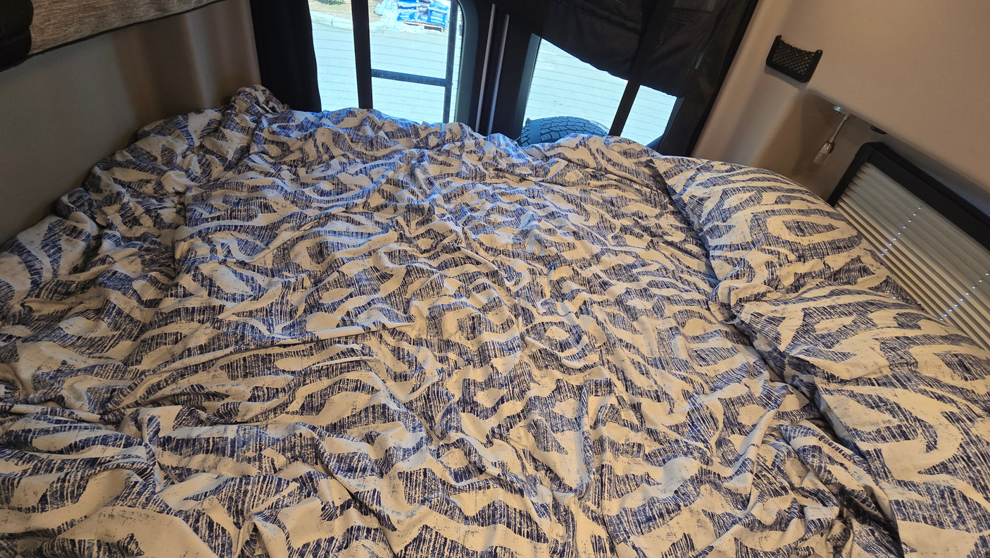 RV rental bedroom: a cozy bed with blue & white patterned bedding, visible rear windows, and side blinds.
