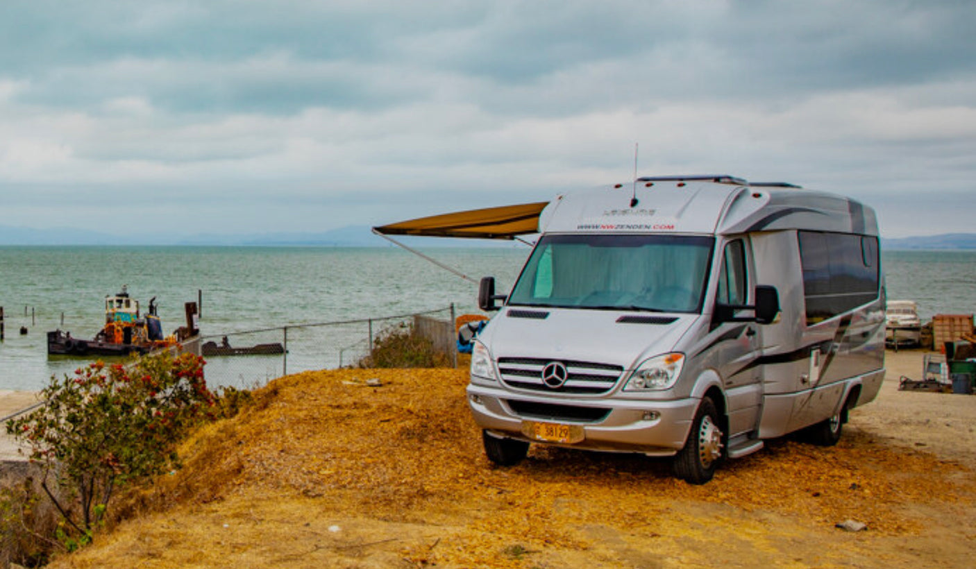 Silver RV rental van with awning extended, parked by the water with an old boat and cloudy sky. Perfect for coastal adventures.