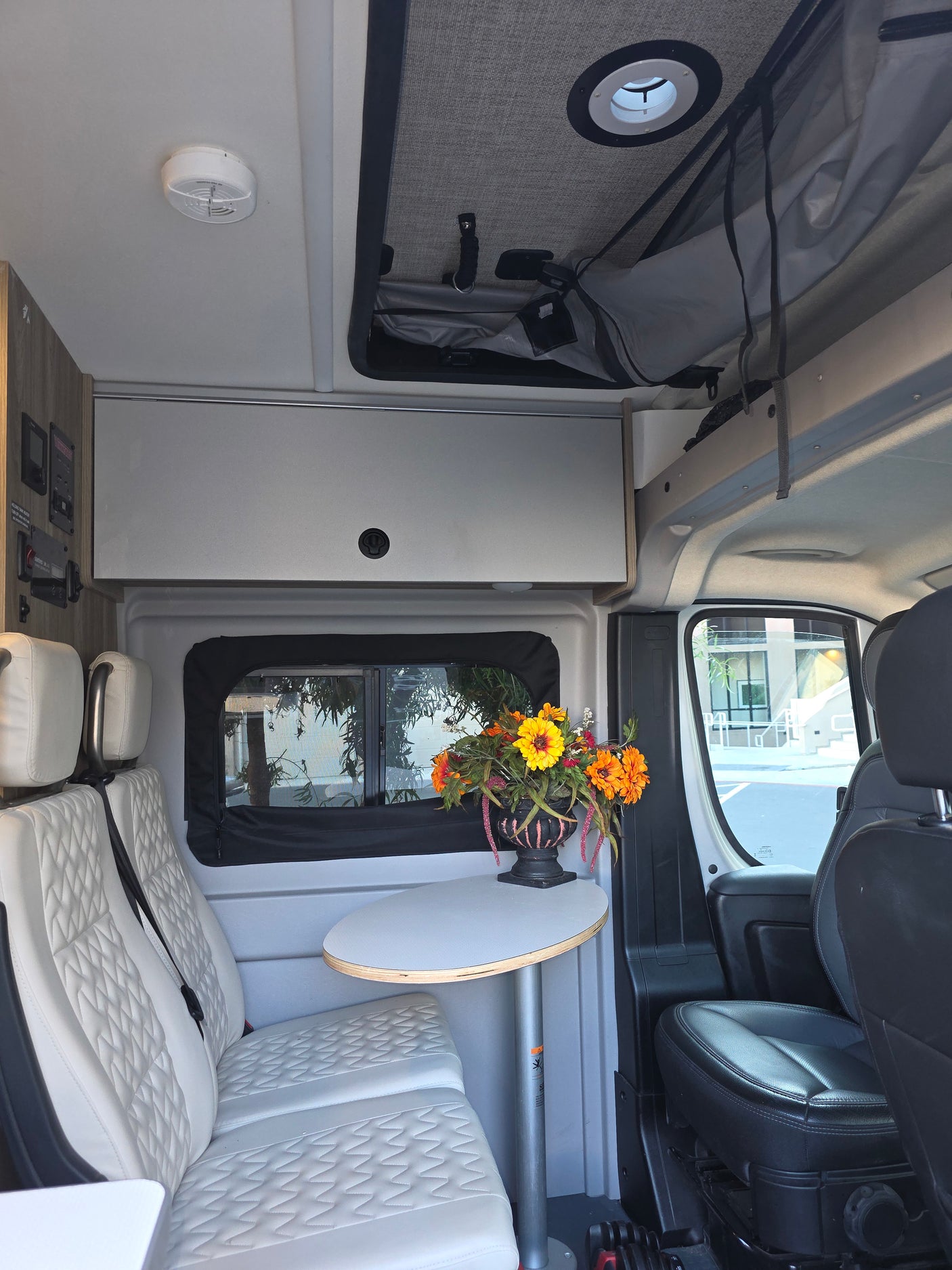 Inside an RV rental: white quilted seats, a round table with flowers, overhead cabinets, and a ceiling vent.