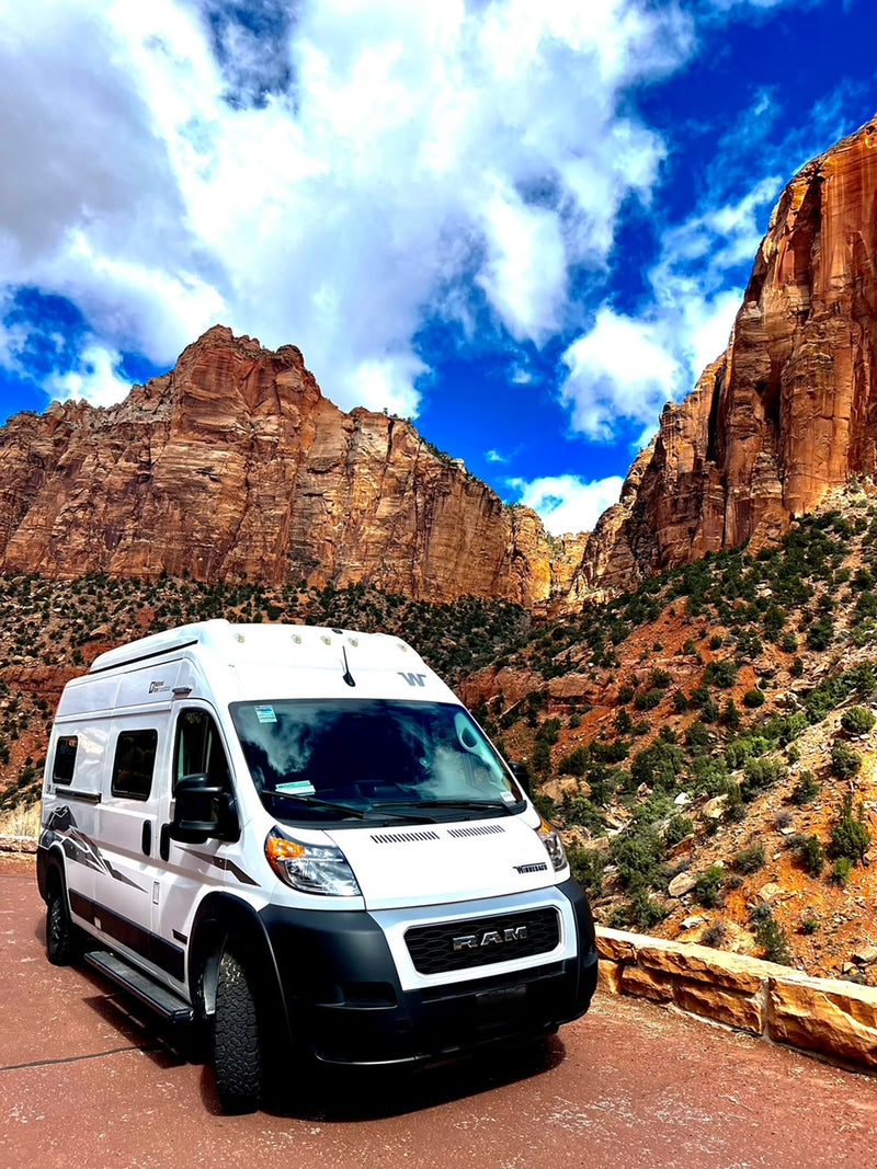 Explore stunning landscapes with an RV rental! White camper van parked by massive red rock cliffs under a dramatic blue sky.|White Winnebago rv rental camper van at a scenic desert campground — photo 1