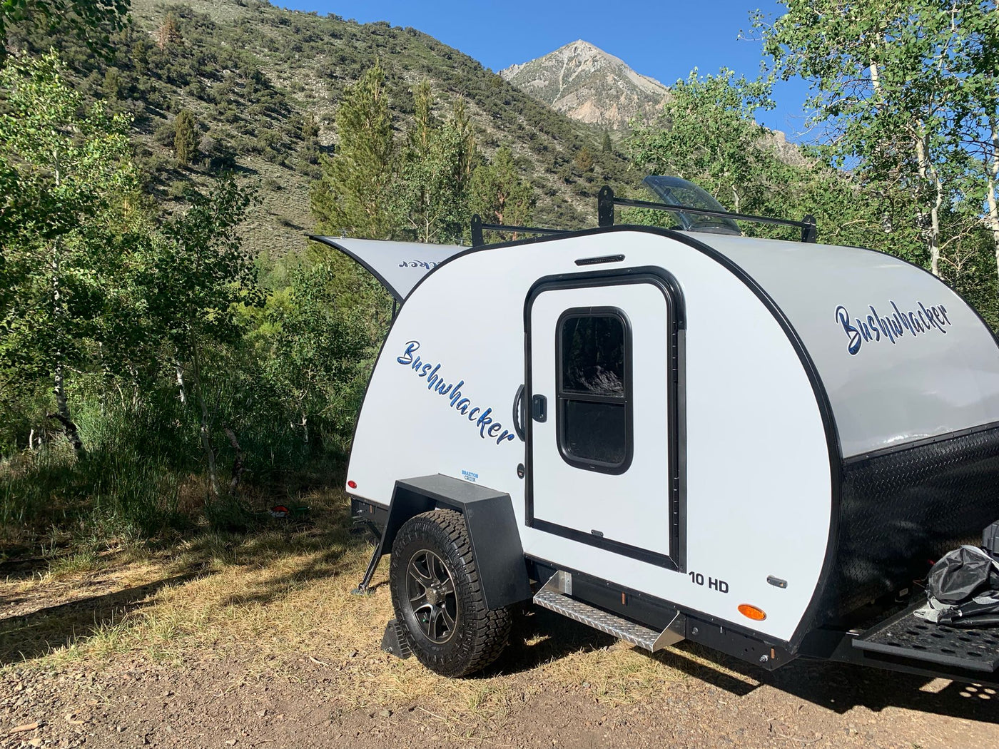  with camping chairs arranged nearby.|Enjoy a scenic RV rental camping trip! This teardrop camper's open kitchen is ready for adventure in the mountains.|Bushwhacker 10 HD teardrop rv rental set up in a beautiful mountain campsite. Enjoy outdoor adventures and nature!|Explore nature with this RV rental: an SUV tows a teardrop trailer amidst green mountains. Perfect for your next camping trip.|RV rental galley kitchen: fully stocked with a K2 cooler — photo 3