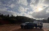 RV rental: Dark SUV towing a white Winnebago trailer on a dirt road, with trees and a cloudy, sun-peeking sky.