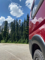 A red RV rental van is parked, foregrounding majestic mountains, evergreen trees, and a cloud-filled sky.