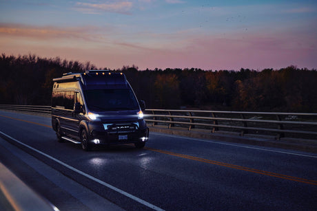 A sleek RV rental camper van, with bright headlights, drives on a bridge at dusk under a vivid pink and blue sky.