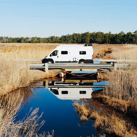 An RV rental, a white camper van, drives over a bridge, its reflection perfectly mirrored in the dark water of a marsh.