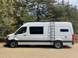White converted camper van, an rv rental, with roof ladder, parked on gravel by trees.