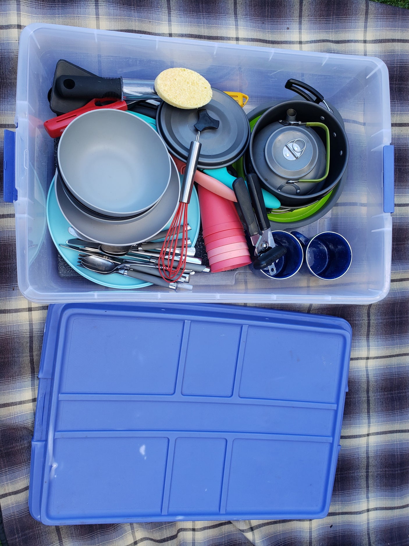 RV rental kitchen kit: A clear bin filled with camping cookware, dinnerware, and utensils on a plaid blanket.