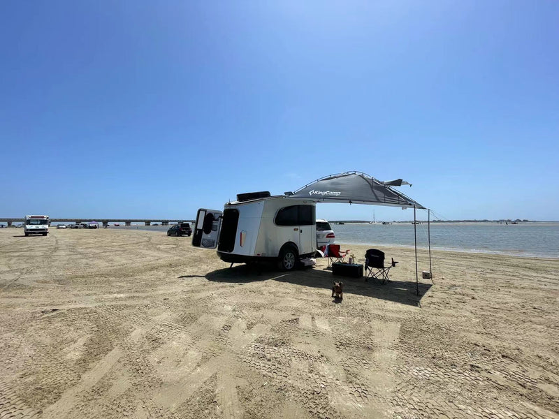 A white tear-drop RV rental parked on a sunny beach with an open side — photo 1