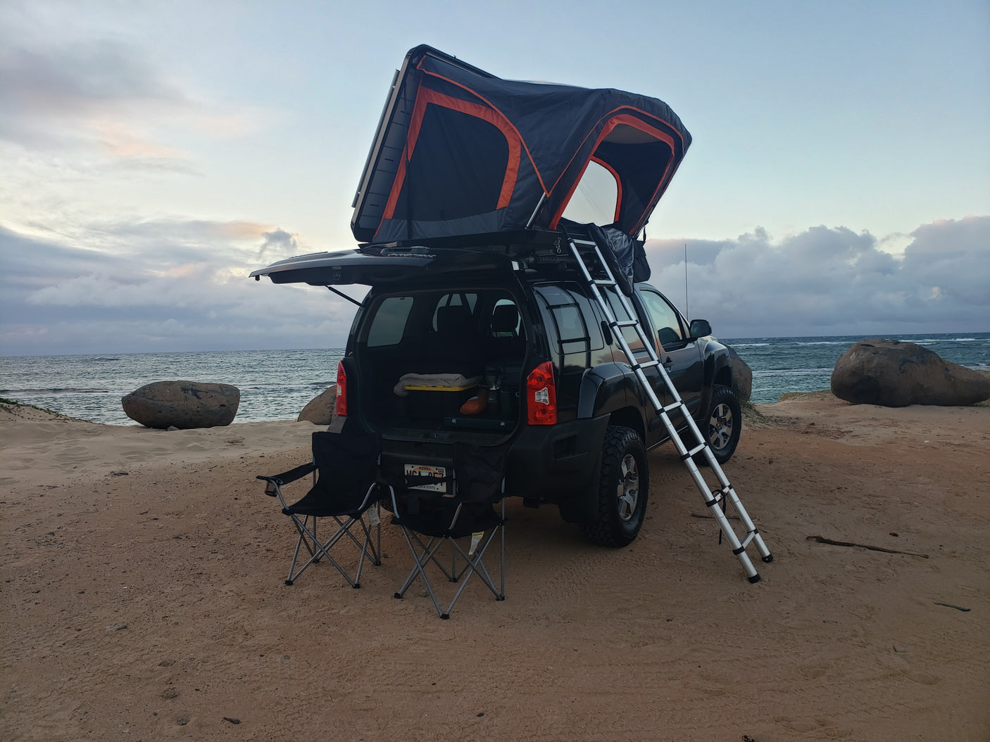 A dark SUV with an open rooftop tent and ladder, parked on a sandy Hawaii beach, showcasing a compact RV rental setup.