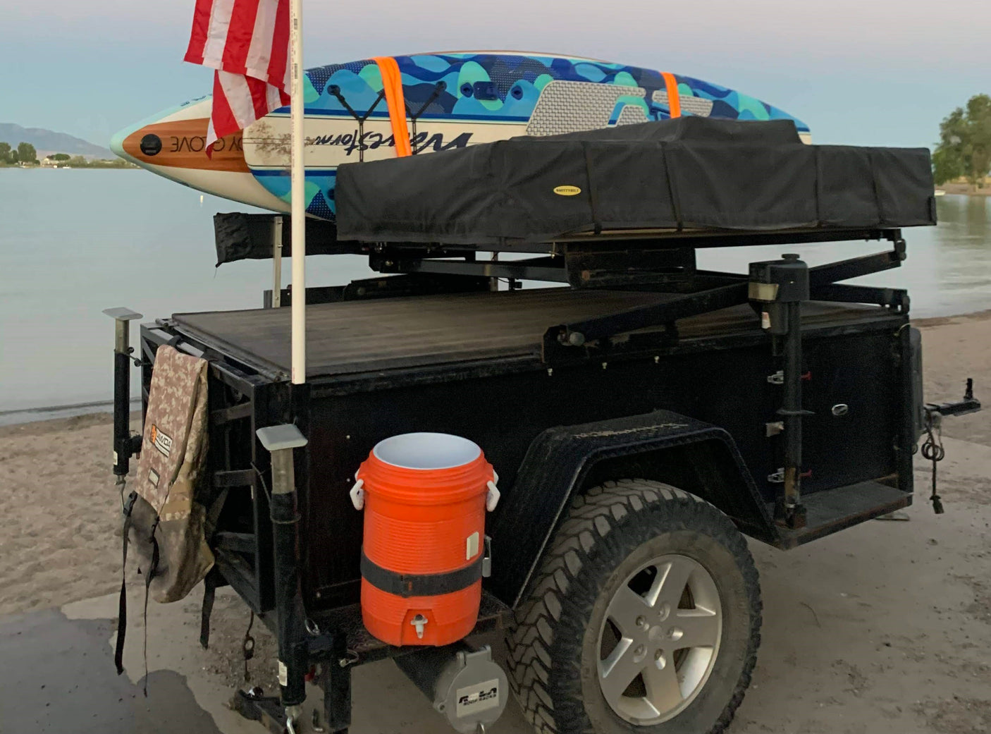 Perfect for your RV rental trip: A utility trailer with rooftop tent, paddleboards, and cooler by the water for outdoor adventures.