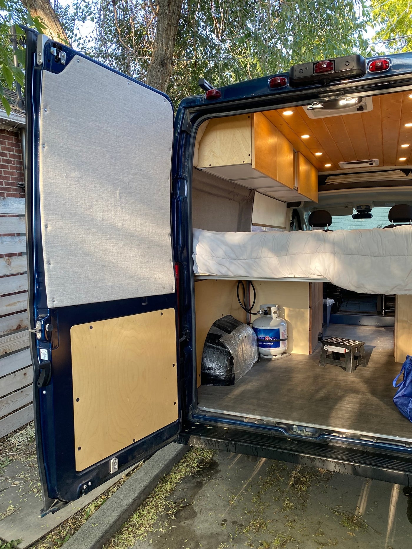 View inside an rv rental camper van, featuring a cozy bed, overhead wooden cabinets, a propane tank, and under-bed storage. — photo 3