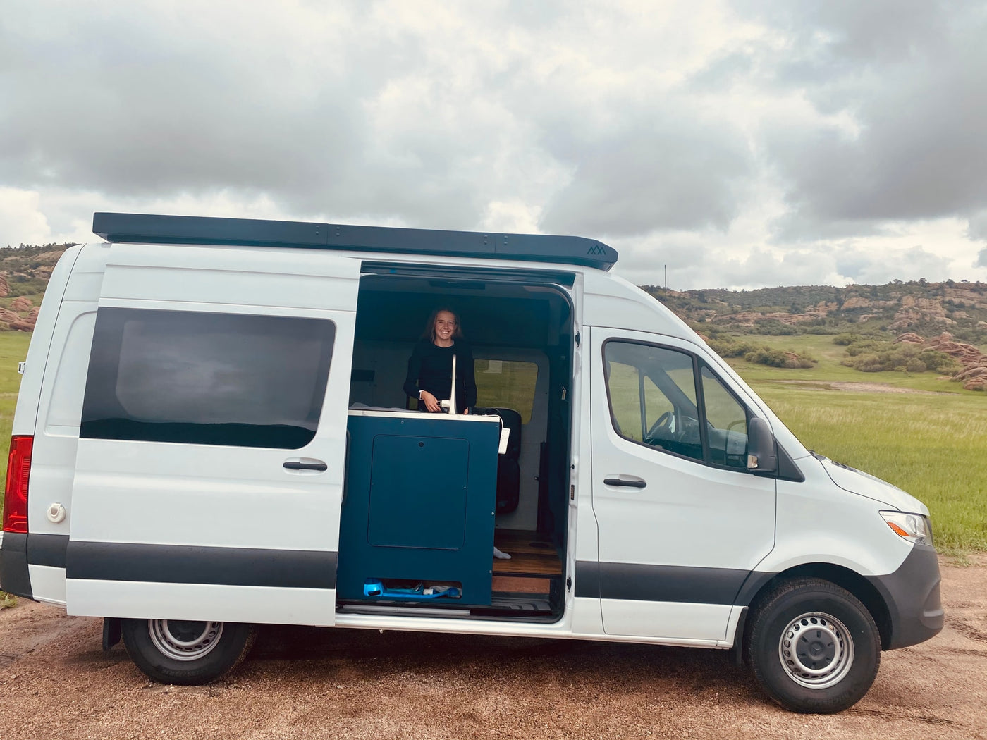 White RV rental van with a smiling woman inside, parked on a dirt road amidst green fields & rocky hills. — photo 7