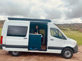 White RV rental van with a smiling woman inside, parked on a dirt road amidst green fields & rocky hills.