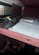 RV rental bunk bed with a light blue blanket, rolled black bag, window, and fire extinguisher below. Ready for adventure.