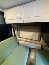 Cozy RV rental interior featuring light green ribbed seating, white overhead storage cabinets, and a window with a shade.