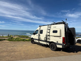 A cream-colored Revel 4x4 RV rental is parked by a scenic ocean coast under a blue sky with wispy clouds.