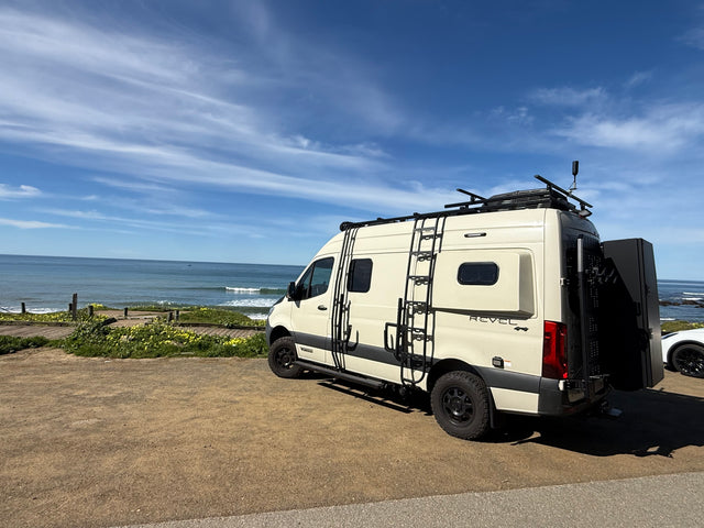 A cream-colored Revel 4x4 RV rental is parked by a scenic ocean coast under a blue sky with wispy clouds.