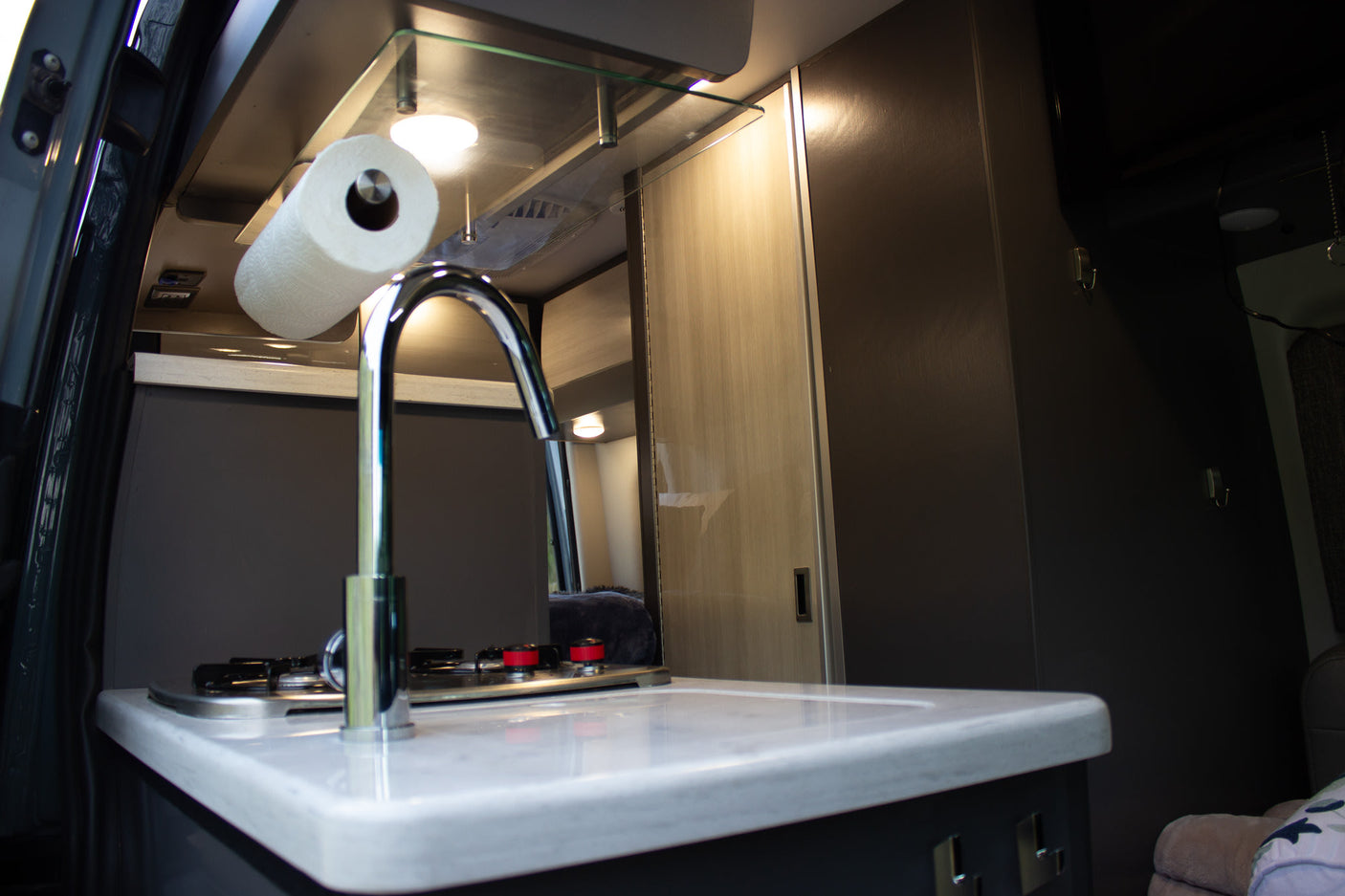 Step inside this RV rental's compact kitchen, featuring a modern sink, stove, white counter, and paper towel holder.