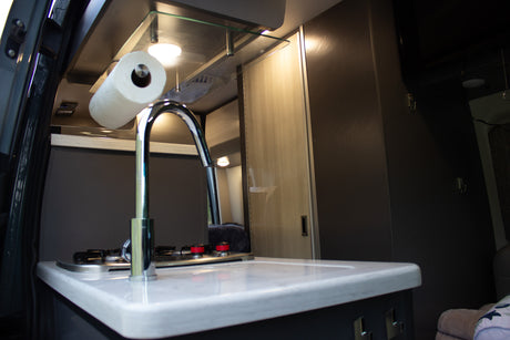 Step inside this RV rental's compact kitchen, featuring a modern sink, stove, white counter, and paper towel holder.