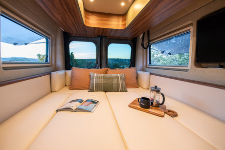 Cozy RV rental interior: lounge bed with coffee, magazine, and scenic views from multiple windows. Perfect for van life adventures!