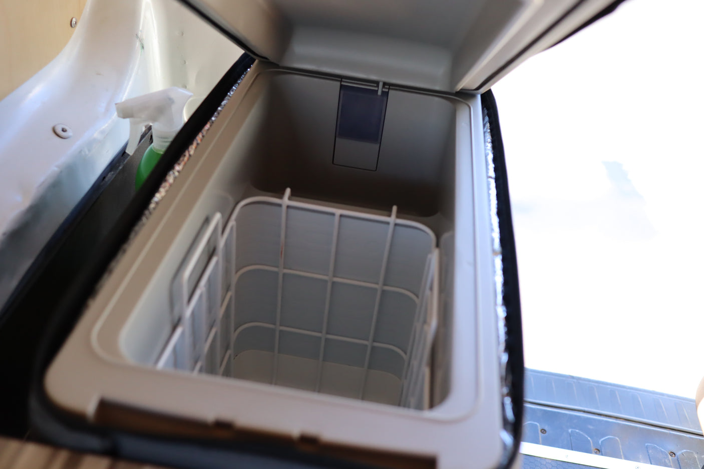 Inside an RV rental's portable cooler, featuring an open lid, interior storage basket, and a spray bottle nearby.