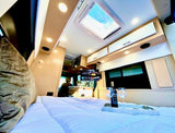 Step inside this luxurious RV rental: a plush bed with wine, TV, skylight, and overhead lighting for comfort and style.