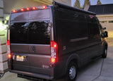 Dark gray camper van, possibly an RV rental, parked in a driveway with its tail lights on at dusk.