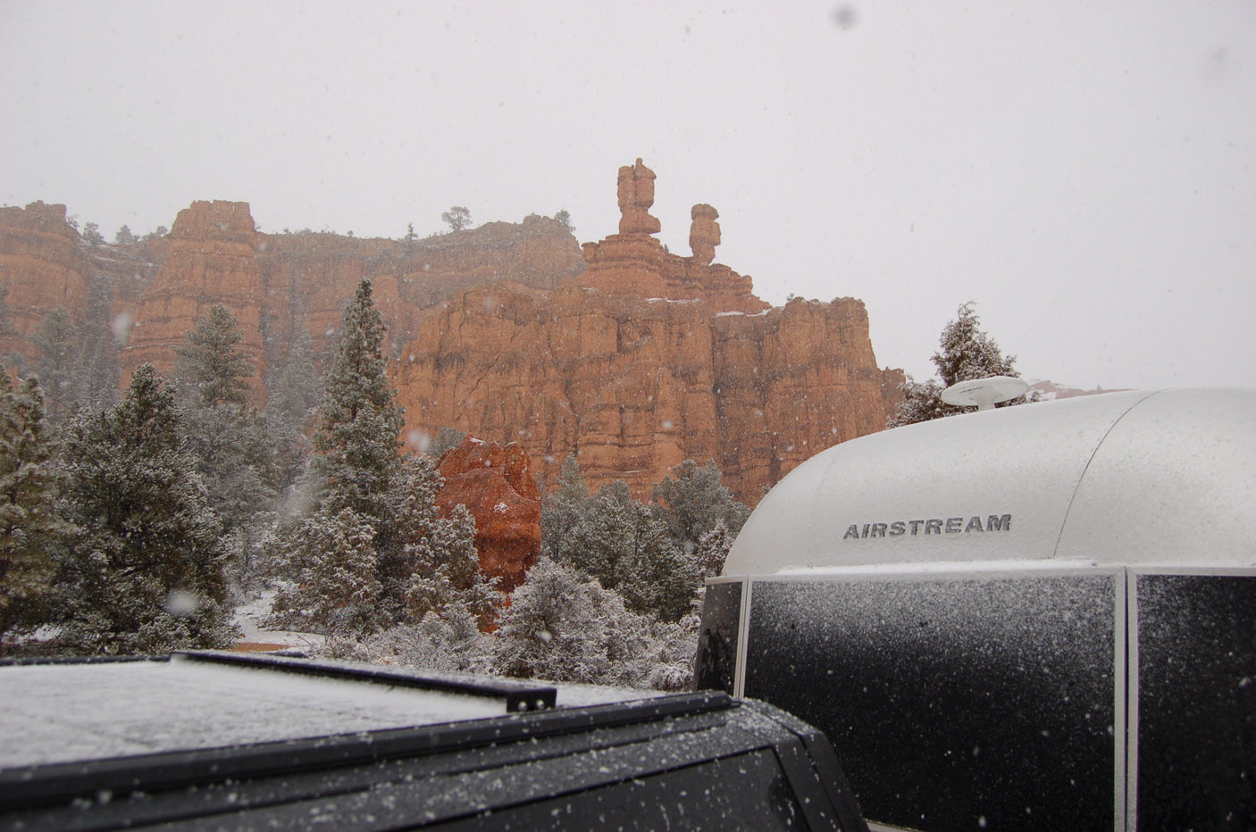 Snow falls on red rock formations, pine trees, and an Airstream rv rental.