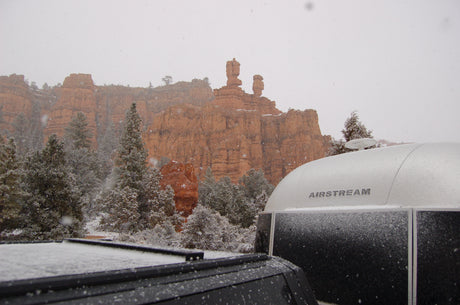Snow falls on red rock formations, pine trees, and an Airstream rv rental.