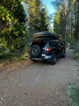 Black SUV with rooftop tent parked in a forest, ideal for an RV rental adventure. Outdoor camp-ready.