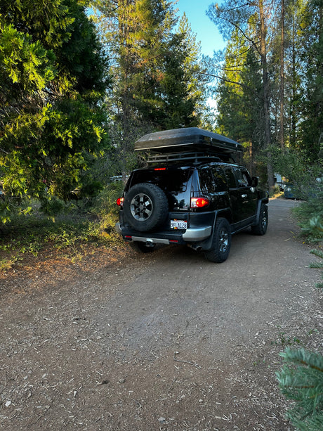 Black SUV with rooftop tent parked in a forest, ideal for an RV rental adventure. Outdoor camp-ready.