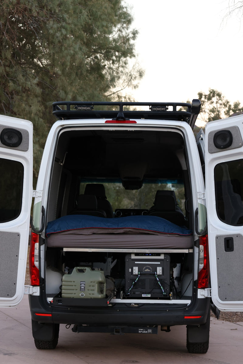 Explore this spacious RV rental camper van with open rear doors, revealing a comfortable bed, portable fridge, and water storage. — photo 11