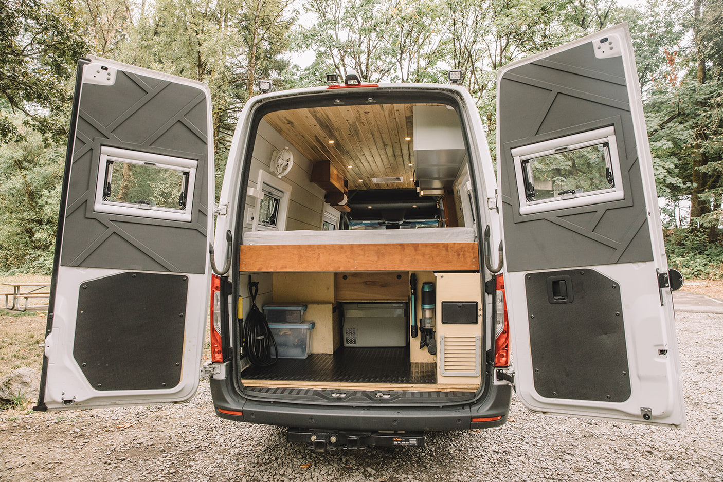 Open rear doors reveal a custom camper van conversion, ideal for rv rental adventures. Features a raised bed, ample storage.