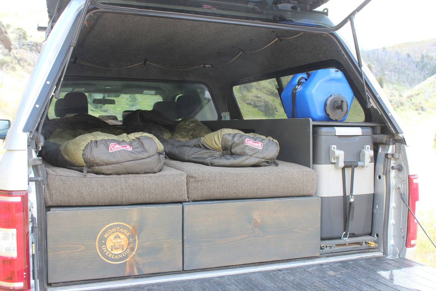 RV rental truck bed camper setup: features a comfortable sleeping area with bags, storage drawers, fridge, and water for overlanding. — photo 7