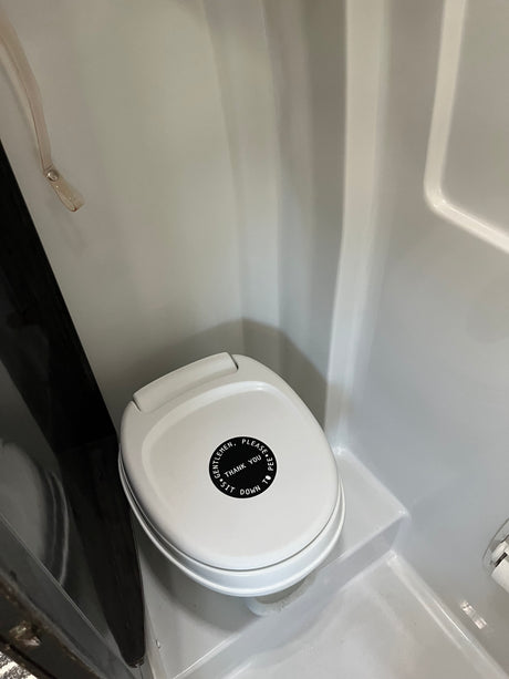 RV rental bathroom features a compact white toilet with a lid sign: 'GENTLEMEN, PLEASE PEE, THANK YOU, SIT DOWN TO'.